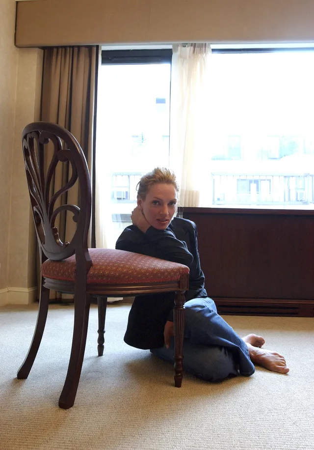 Uma Thurman sitting barefoot indoors in a dark top and jeans.