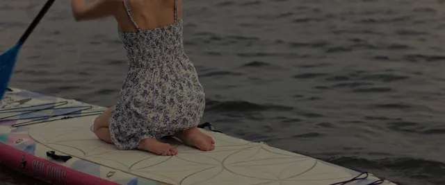 Anna Kendrick kneeling barefoot on a paddleboard wearing a patterned dress.