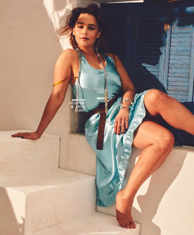 Emilia Clarke sitting barefoot on sunlit steps in a blue dress.