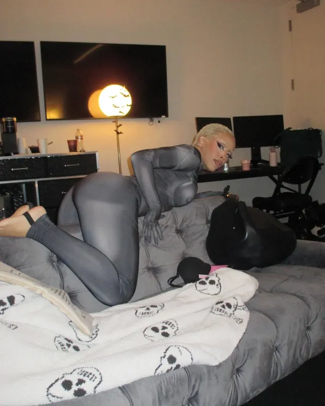 Doja Cat kneeling on a bed in a grey outfit with her bare soles visible.