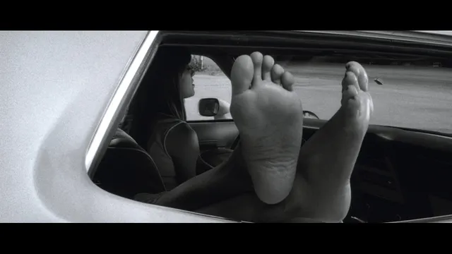 Rosario Dawson resting her feet outside a car window in a monochrome screen image.