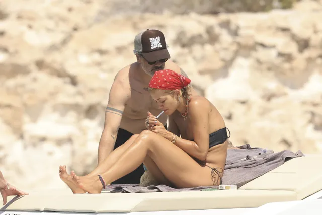 Rita Ora sitting barefoot on a beach lounger.