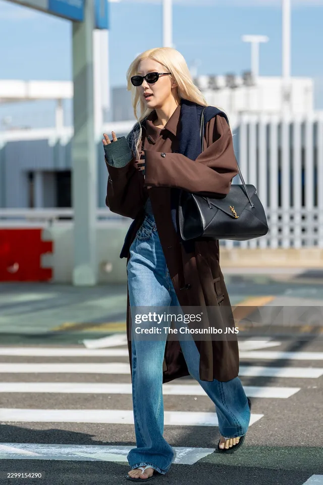 Rosé crossing a street in a brown coat and blue jeans with open-toe sandals.