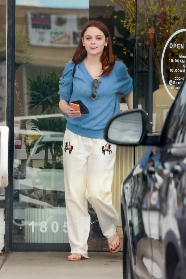 Joey King walking outside in a blue top, pale trousers and flat sandals.
