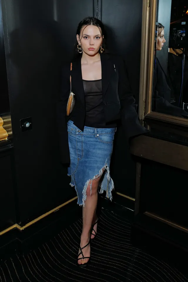 Dafne Keen standing indoors in a jacket and denim skirt with heels.