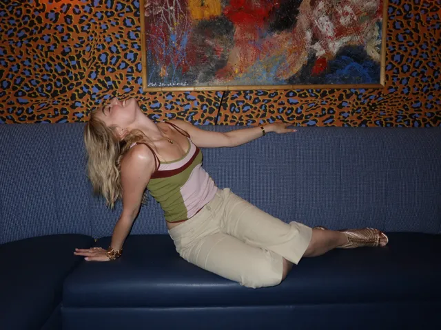 Meg Donnelly reclining barefoot on a blue sofa in a colorful top and pale skirt.