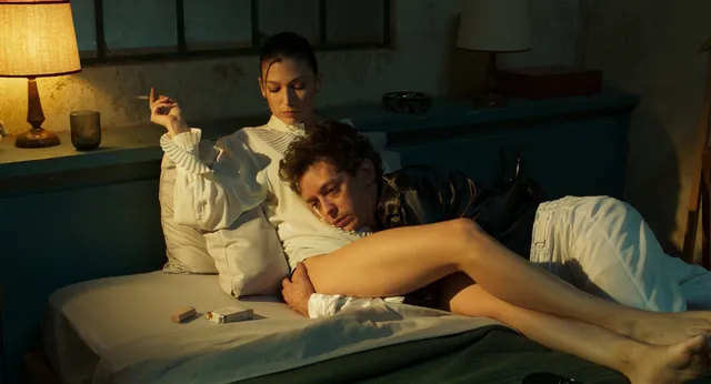 Úrsula Corberó reclining barefoot beside a man on a bed in a scene.