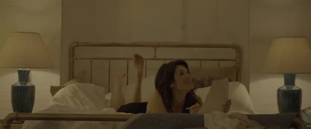 Gemma Arterton sitting in bed in a softly lit screen scene.