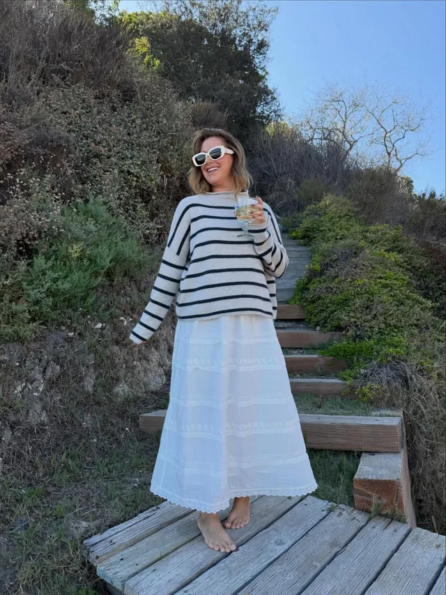 Ashley Tisdale standing barefoot on a wooden terrace in a striped top and long pale skirt.