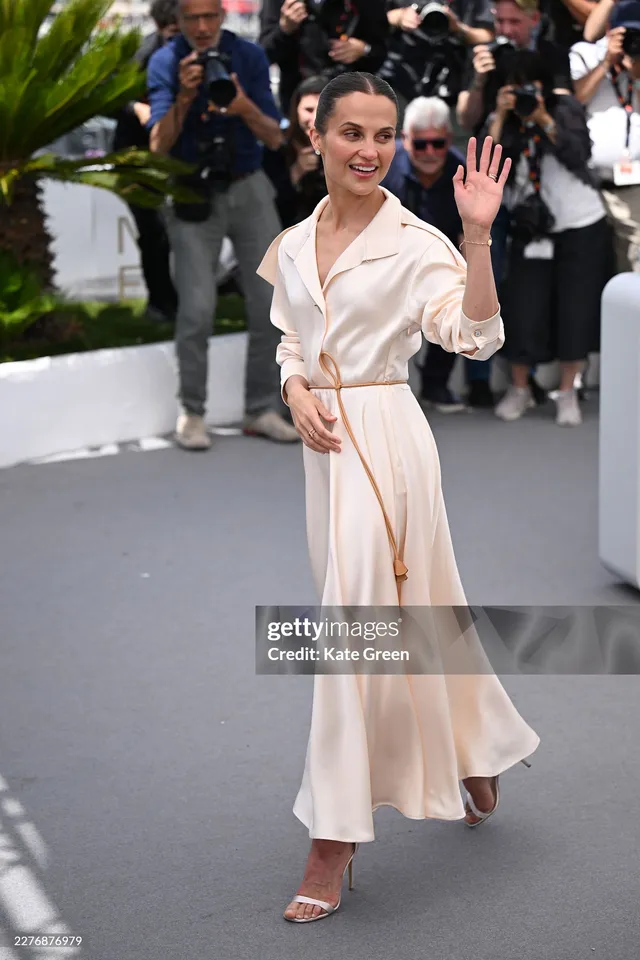 Alicia Vikander waving to photographers in a cream skirt.
