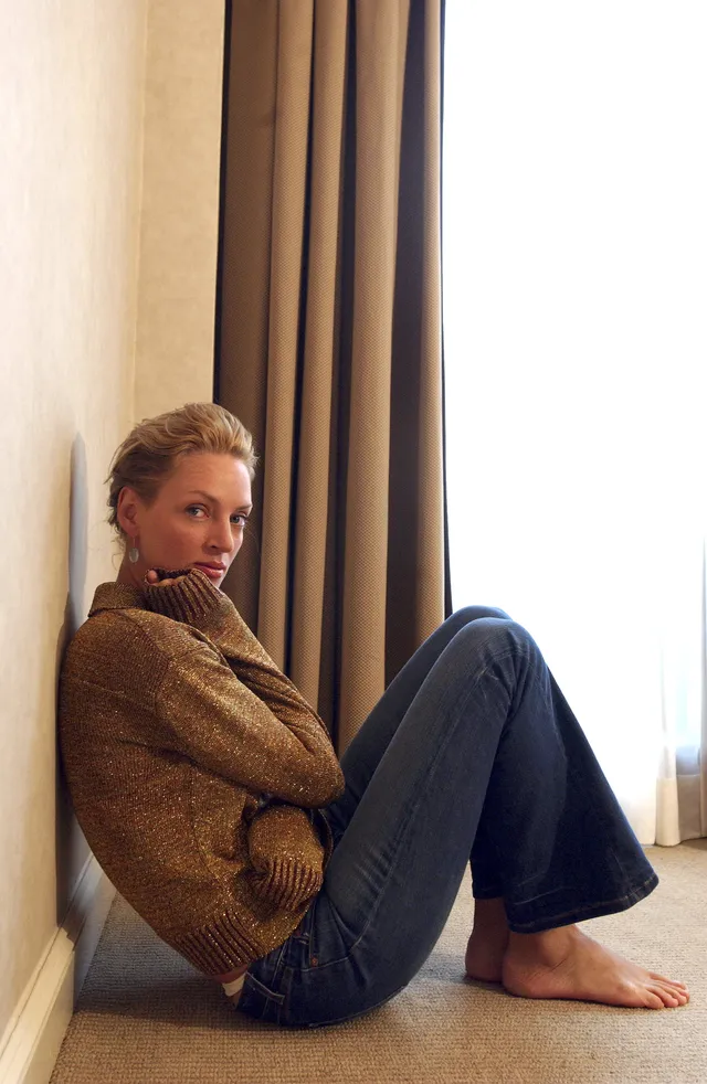Uma Thurman sitting barefoot against a wall in jeans.