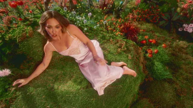Hilary Duff lying barefoot on grass among flowers in a pink dress.