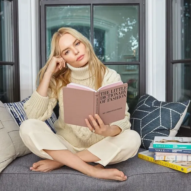 Kate Bosworth sitting cross legged on a sofa reading a book.