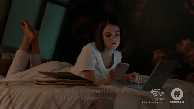 Maia Mitchell lying barefoot on a bed with a laptop.
