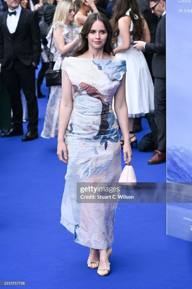 Felicity Jones walking on a blue carpet in a printed dress.
