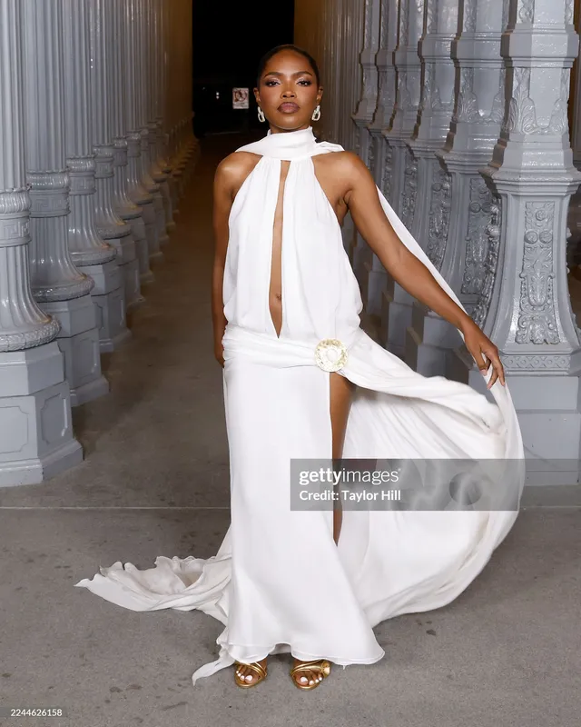 Ryan Destiny lifting the hem of a white gown while wearing gold heels.