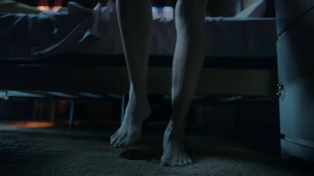 Peyton List standing barefoot beside furniture in a dark room.