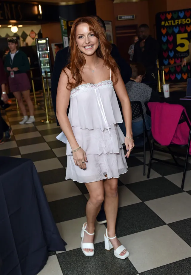 Mckenna Grace standing indoors in a short white dress and heels.