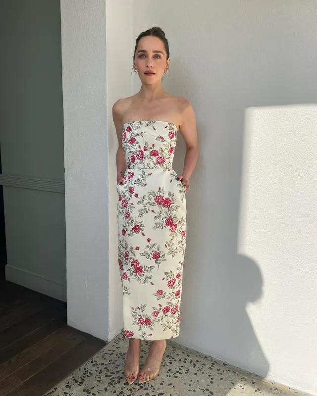 Emilia Clarke standing in a floral dress and heels beside a wall.