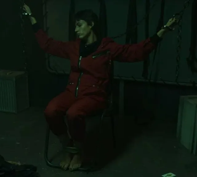 Úrsula Corberó sitting barefoot in a red jumpsuit in a dimly lit scene.