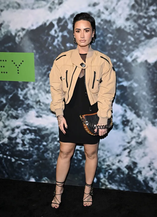 Demi Lovato posing at an event in a tan jacket, short skirt and heels.