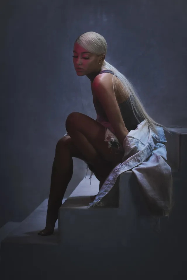 Ariana Grande seated barefoot on a bed in a pale dress for a portrait.