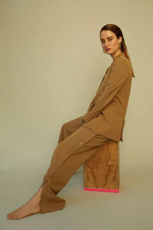Grace Van Patten sitting barefoot on a wooden block in a brown suit.