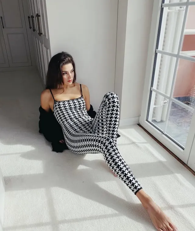 Hande Erçel sitting barefoot by a window in a checked outfit.
