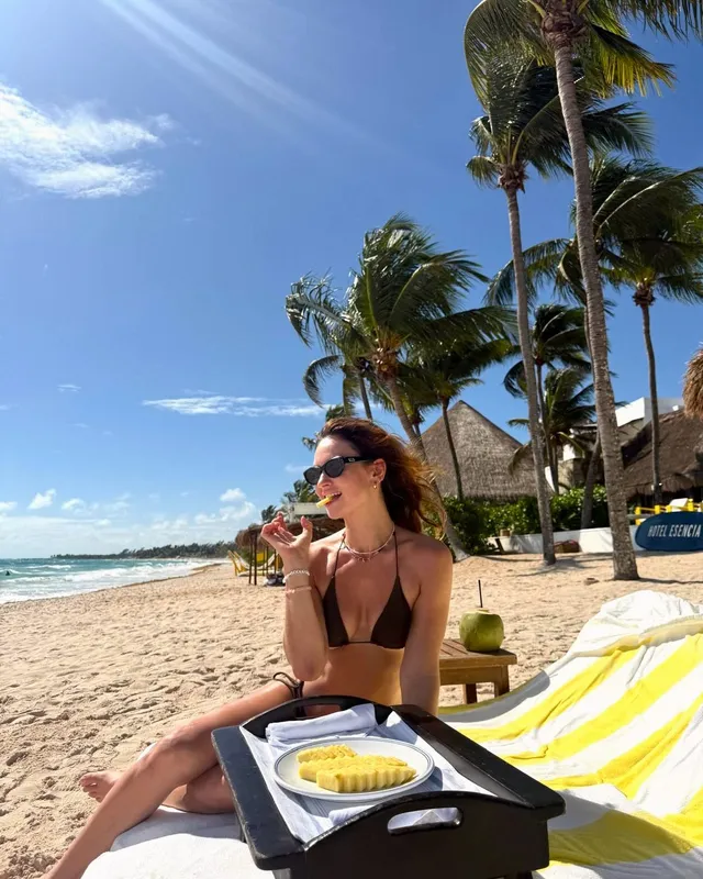 Lily James sitting barefoot on a tropical beach lounger.
