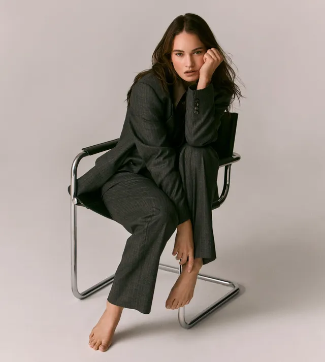 Lily James sitting barefoot on a chair in a dark suit.