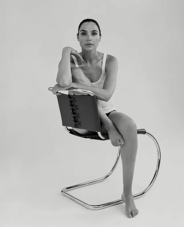 Gal Gadot sitting barefoot on a chair for a monochrome portrait.