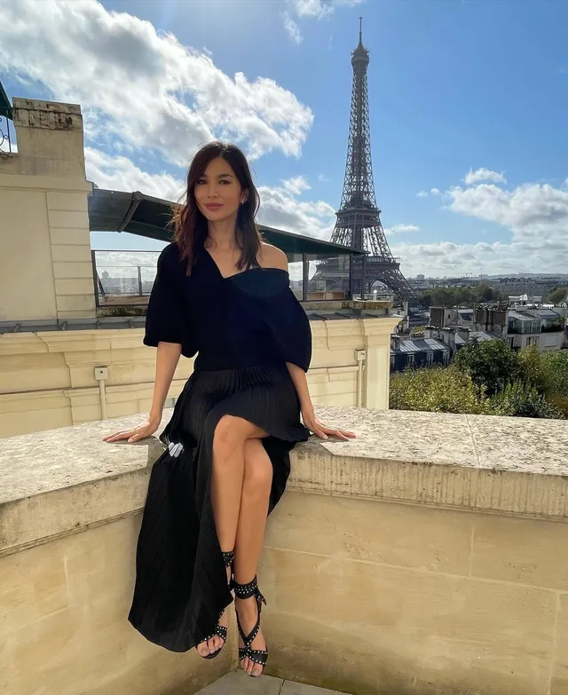 Gemma Chan sitting on a stone ledge near the Eiffel Tower.