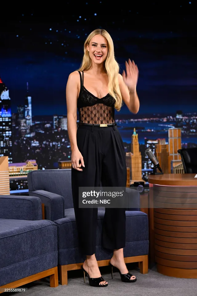 Grace Van Patten waving on a television set in a black outfit and heels.