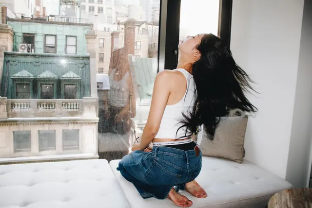 Jennie Kim kneeling barefoot on a window seat in jeans and a white top.