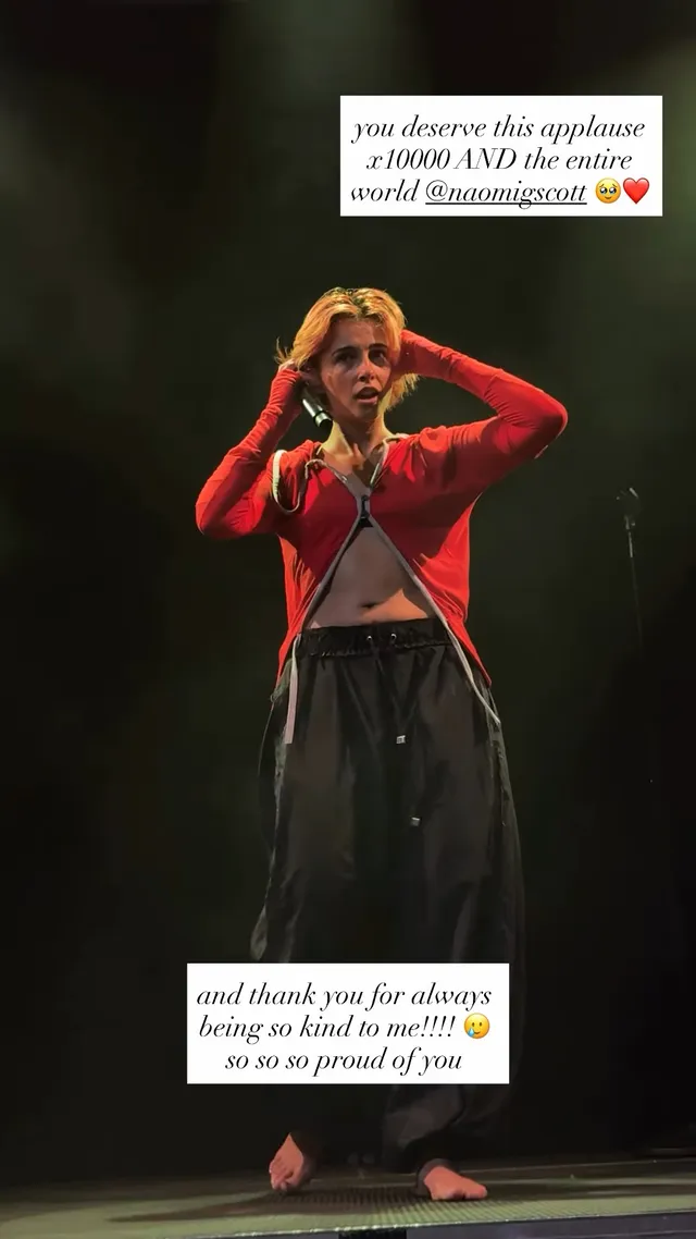 Naomi Scott singing barefoot in a red jacket onstage.