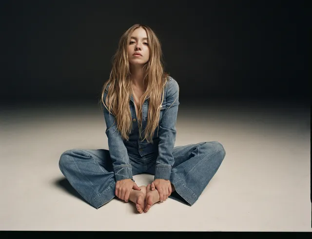 Sydney Sweeney: seated cross-legged and barefoot against a dark background.