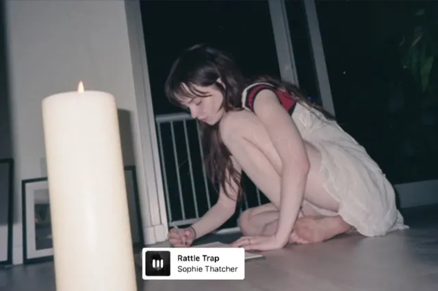 Sophie Thatcher crouching barefoot beside a large candle indoors.