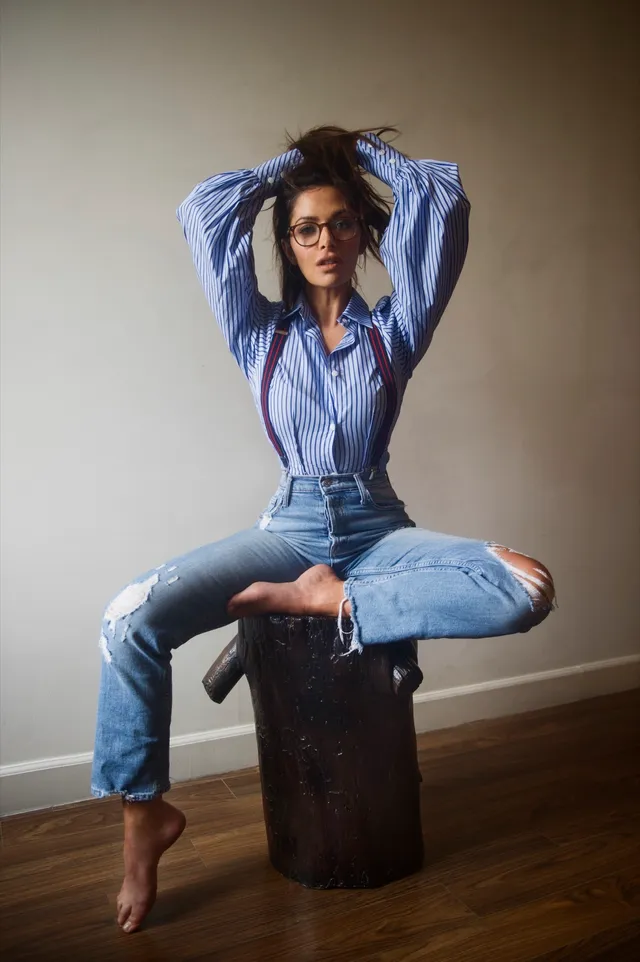 Sarah Shahi sitting on a wooden stool in a denim outfit.