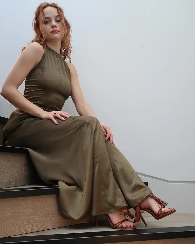 Joey King sitting on a studio bench in an olive dress and brown heels.