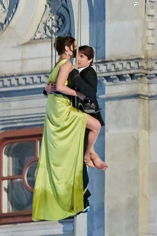 Rebecca Ferguson posing barefoot in a yellow dress on a rooftop railing beside a companion.