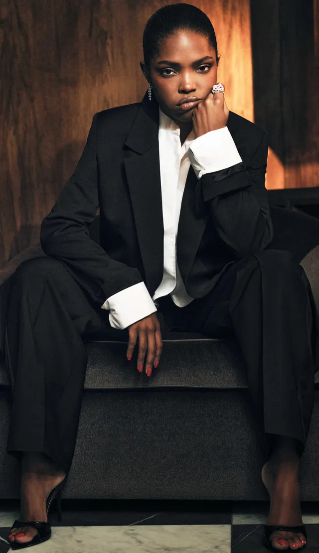 Ryan Destiny sitting in a black suit and black heels for a portrait.