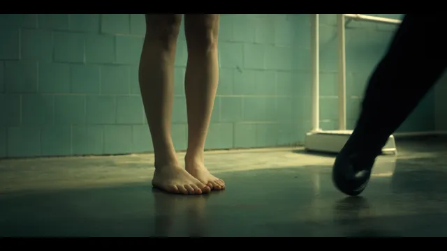 Grace Van Patten standing barefoot beside a tiled wall in a film scene.
