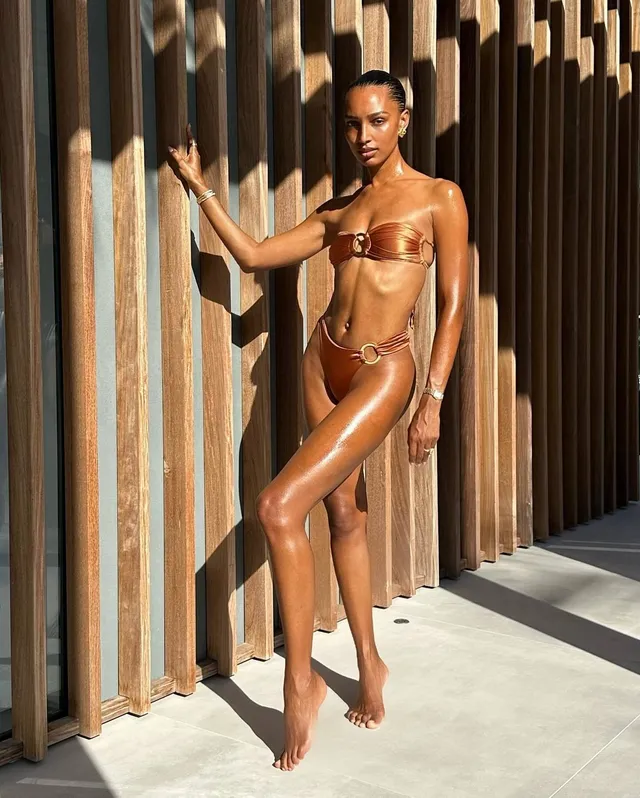 Jasmine Tookes standing barefoot beside a wooden wall in metallic swimwear.