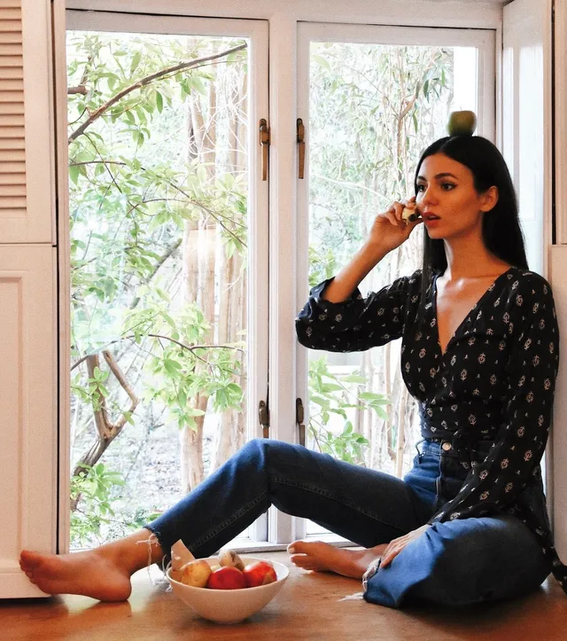 Victoria Justice sitting barefoot beside a window wearing jeans.