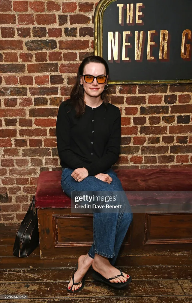 Daisy Ridley sitting in a pub in a black top and jeans.