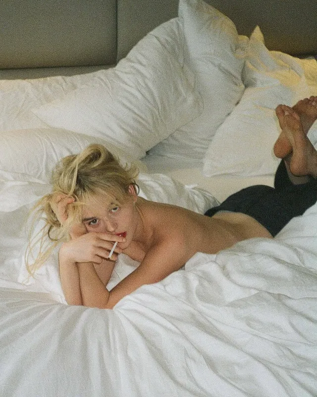 Emily Alyn Lind reclining on a white bed with bare feet visible.