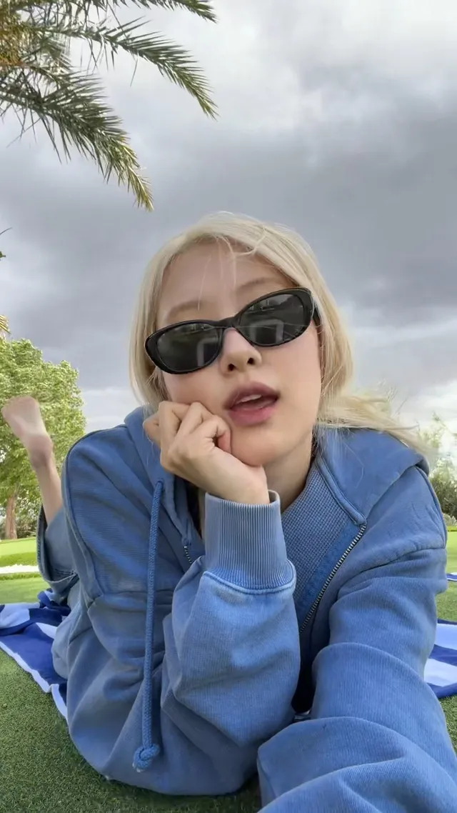 Rosé resting her chin on her hand while seated outside in a blue jacket and sunglasses.