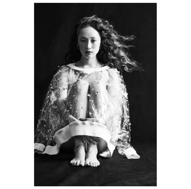 Havana Rose Liu crouching barefoot in a pale dress in a monochrome photograph.