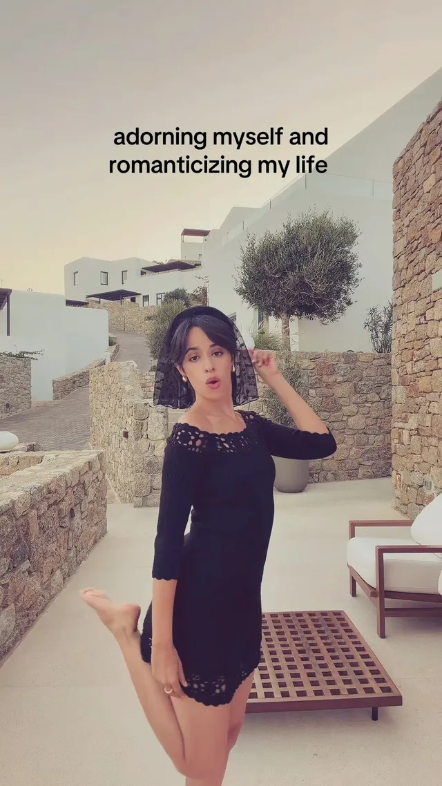 Camila Cabello posing outdoors in a dark outfit with a bare foot raised.
