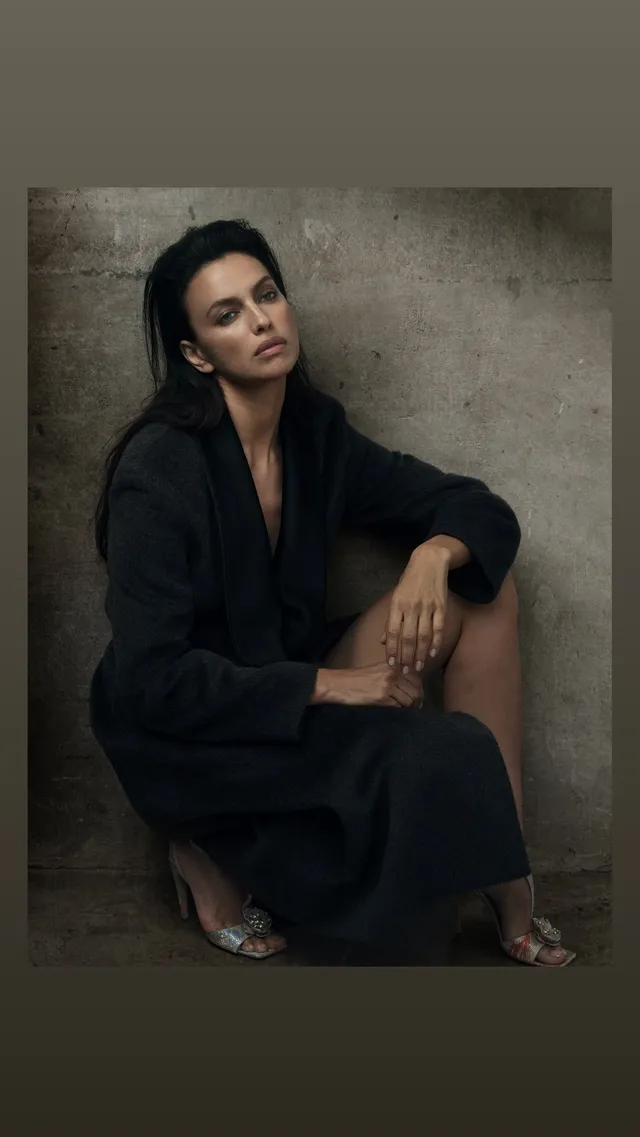 Irina Shayk sitting in a black outfit and heels for a studio portrait.